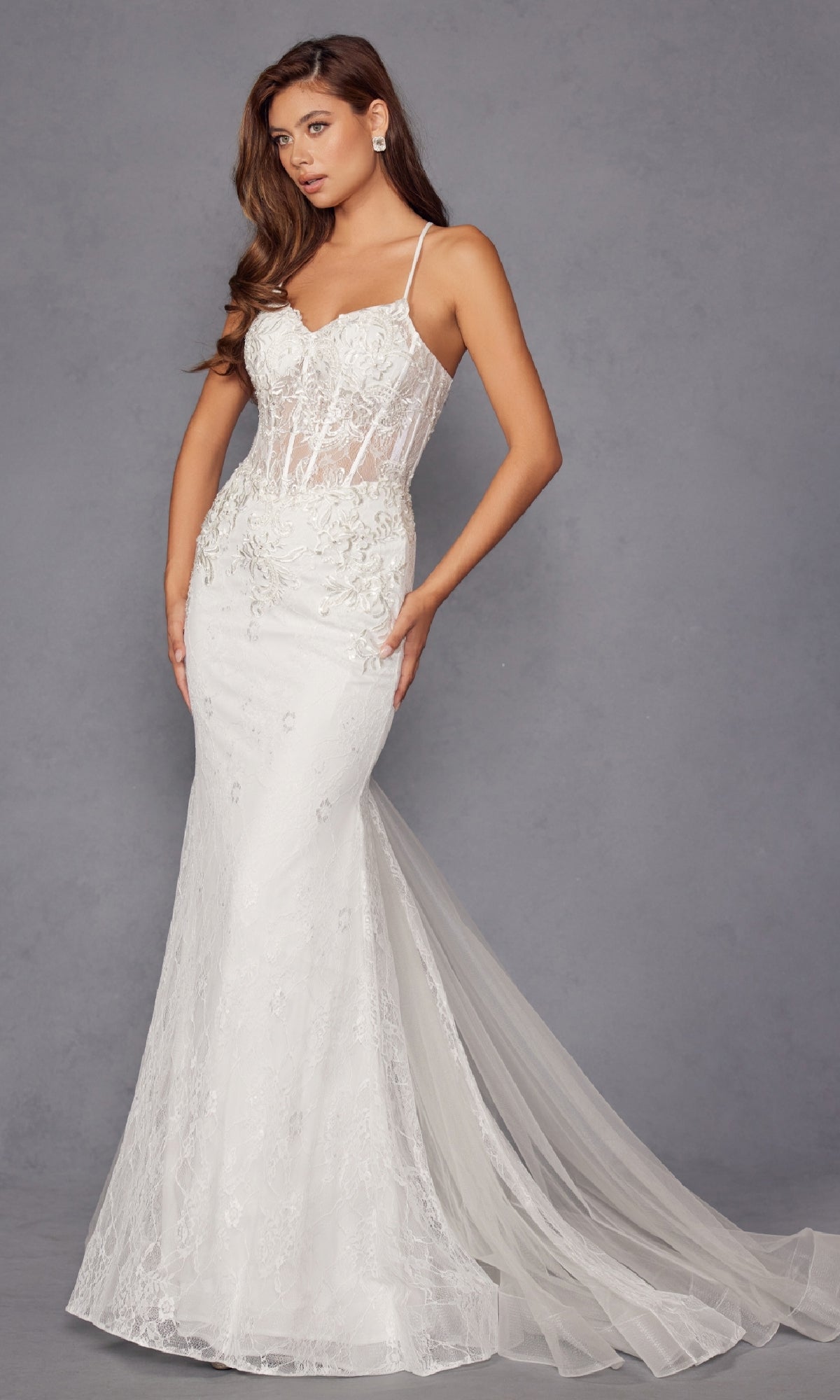 A woman models the Long White Bridal Dress: Juliet JT2481UW, featuring floral lace embroidery, a sheer corset bodice, thin straps, and a long tulle train, standing against a plain gray background.
