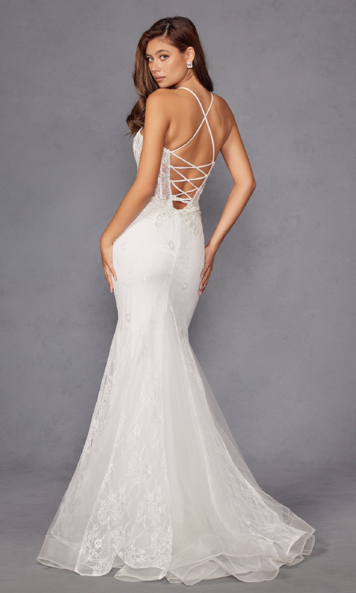 A woman stands against a neutral gray background wearing the Long White Bridal Dress: Juliet JT2481UW, featuring an off-white lace design with a flared hem, sheer corset bodice, and floral lace embroidery.