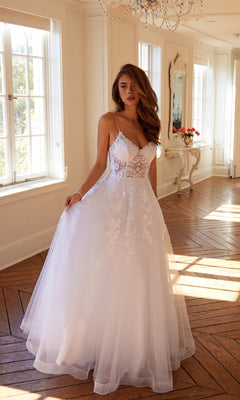 A woman in the Long Off-White Prom Dress: Juliet JT2482SW stands in a bright room with large windows, wooden floors, and elegant decor.