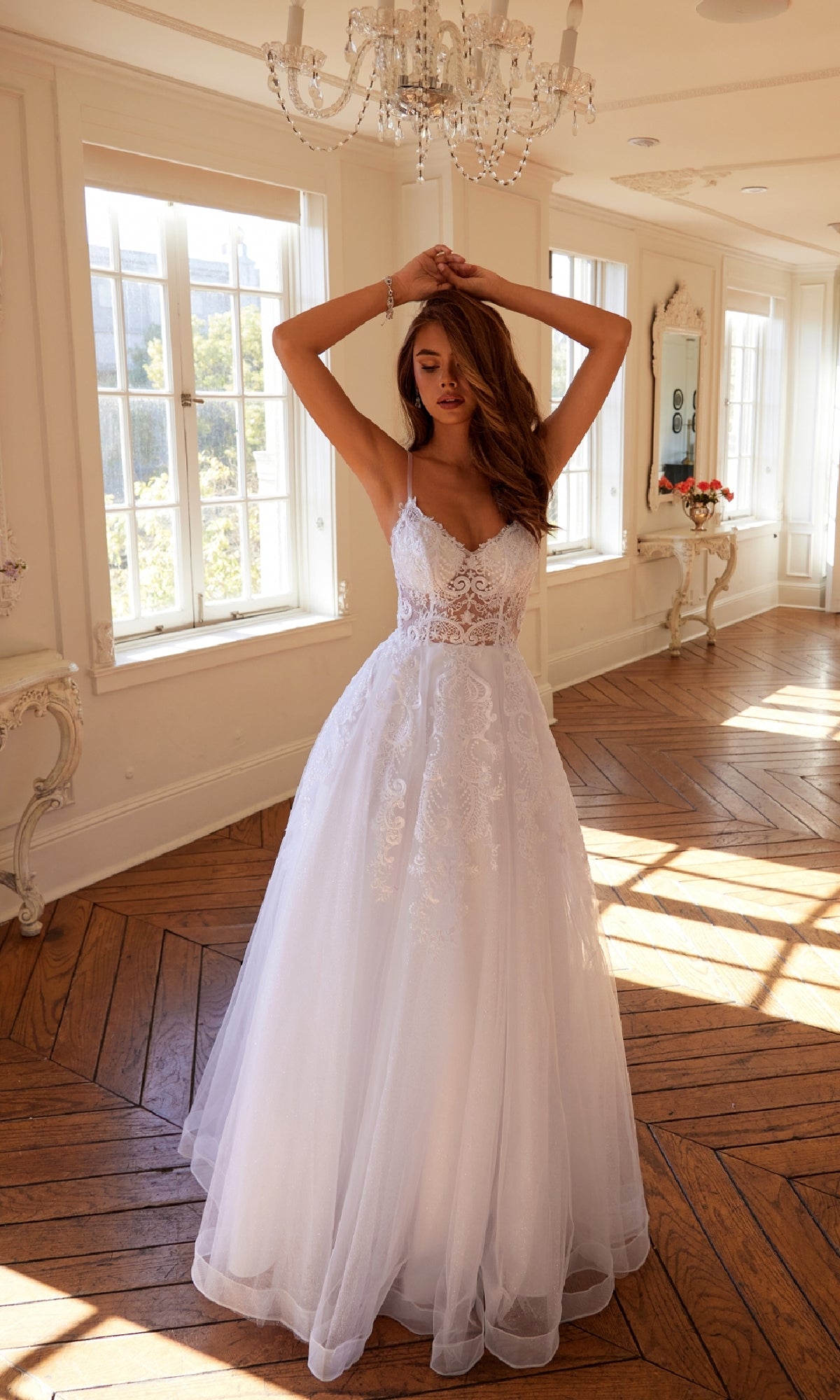 A woman wearing the Long Off-White Prom Dress: Juliet JT2482SW stands in a sunlit room with large windows, hardwood floors, and elegant decor, raising her arms above her head.