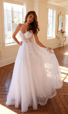 A woman stands in a sunlit room with wooden floors and large windows, wearing the Juliet JT2482SW Long Off-White Prom Dress featuring a lace bodice, tulle A-line skirt, and thin straps.