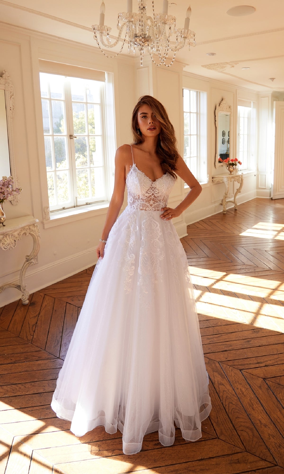 A woman wearing the Long Off-White Prom Dress: Juliet JT2482SW stands in a sunlit room with large windows, wooden floors, and ornate white furniture.