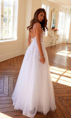 A woman in the Long Off-White Prom Dress: Juliet JT2482SW with a lace bodice and sleeveless design stands on a wooden floor in a sunlit room with large windows.