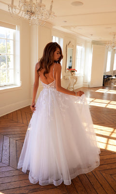 A woman wears the Juliet JT2482SW long off-white prom dress, featuring lace details and a tulle A-line skirt, as she stands in a bright room with large windows, wooden floors, and chandeliers.