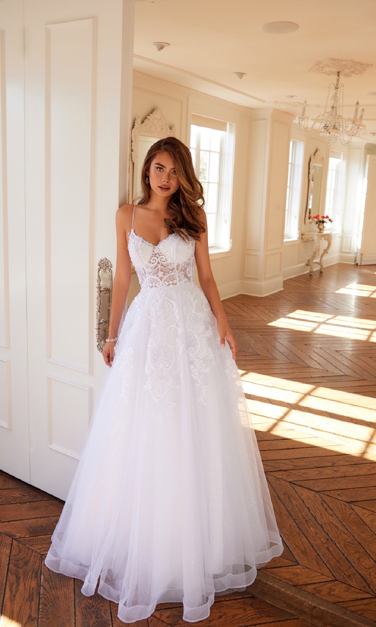 A woman wears the Long Off-White Prom Dress: Juliet JT2482SW, standing in a bright room with large windows, wooden floors, and chandeliers.