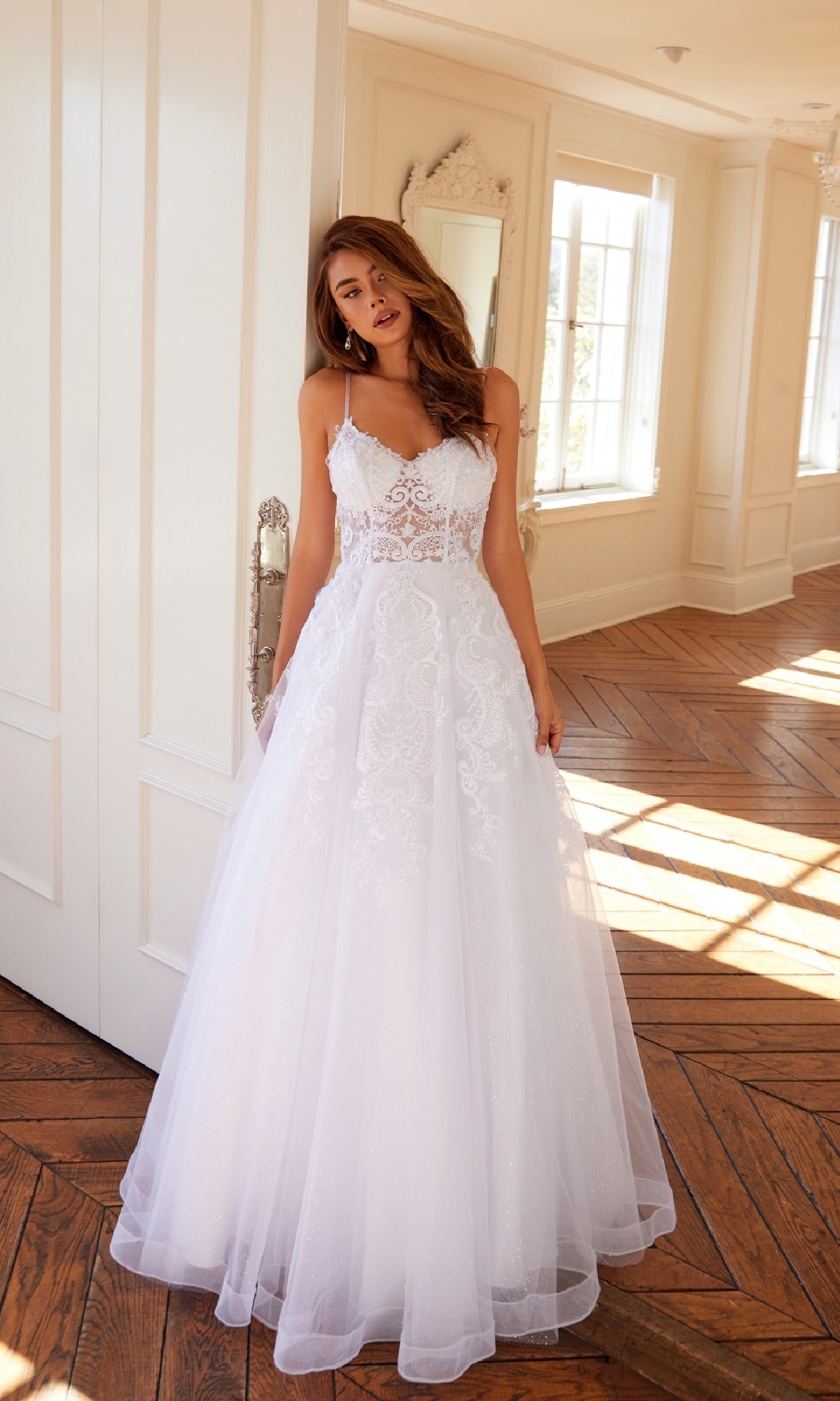 Wearing the Long Off-White Prom Dress: Juliet JT2482SW, a woman stands indoors on a wooden floor, leaning against a white door in a bright room with large windows.