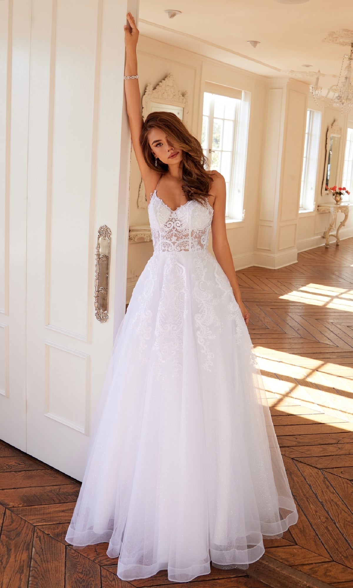 A woman in the Long Off-White Prom Dress Juliet JT2482SW, featuring a lace bodice and tulle A-line skirt, stands indoors with one arm raised against a door in a sunlit room.