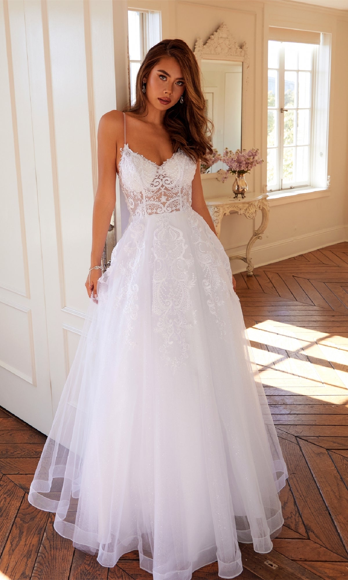 A woman stands indoors wearing the Long Off-White Prom Dress: Juliet JT2482SW, a sleeveless lace gown with a tulle A-line skirt, in a room with large windows and wooden floors.