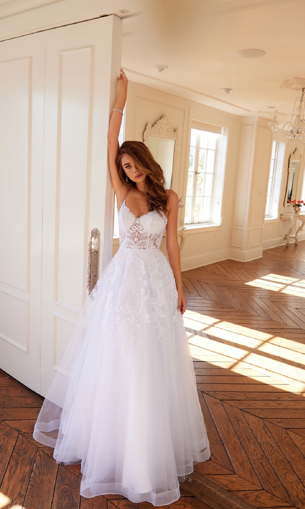 A woman wearing the Long Off-White Prom Dress: Juliet JT2482SW stands indoors, resting an arm on a door as sunlight pours in through large windows.