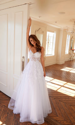A woman wearing the Long Off-White Prom Dress: Juliet JT2482SW stands indoors, resting an arm on a door as sunlight pours in through large windows.