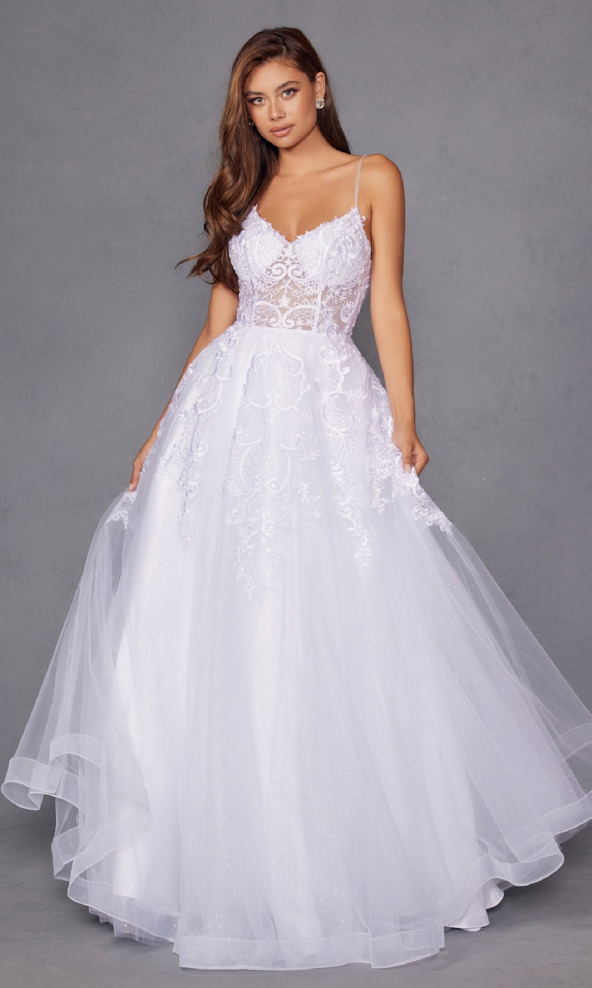 A woman wearing the Long Off-White Prom Dress: Juliet JT2482SW, featuring a delicate lace bodice and tulle A-line skirt, stands against a gray background, holding the sides of her gown.