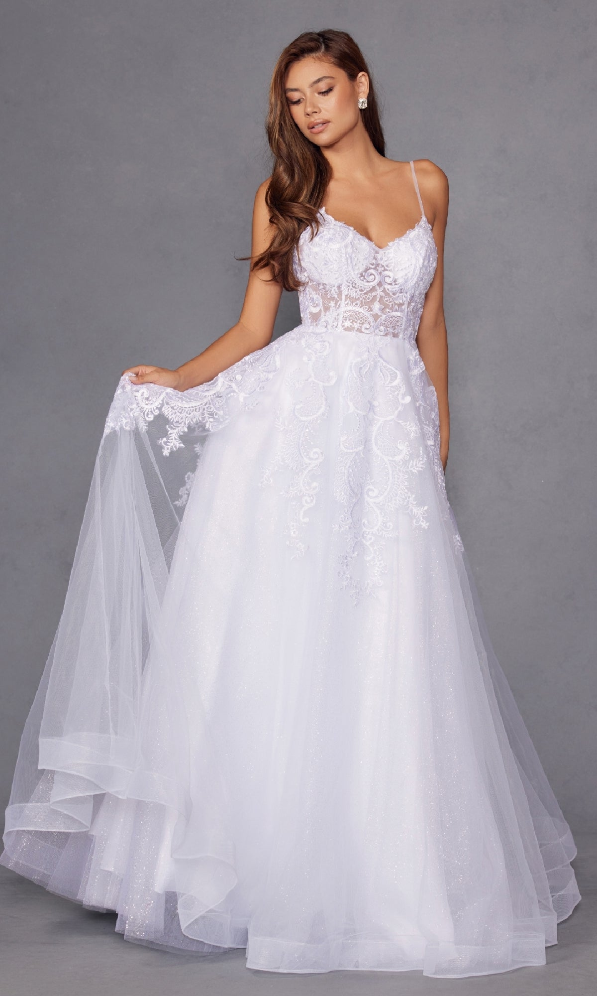 A woman models the Juliet JT2482SW long off-white prom dress with a lace bodice, thin straps, flowing tulle A-line skirt, floral embroidery, and sheer details, standing against a plain gray background.