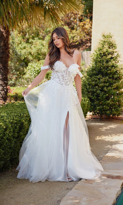A woman stands outdoors near lush greenery wearing the Off-White Bridal Ball Gown: Juliet JT2483KW, featuring an off-the-shoulder design, floral details, a sheer skirt, and an embroidered corset bodice.