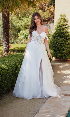 A woman stands in a garden wearing the Off-White Bridal Ball Gown: Juliet JT2483KW, featuring an off-the-shoulder design, floral details, embroidered corset bodice, and a tulle skirt with a thigh-high slit.