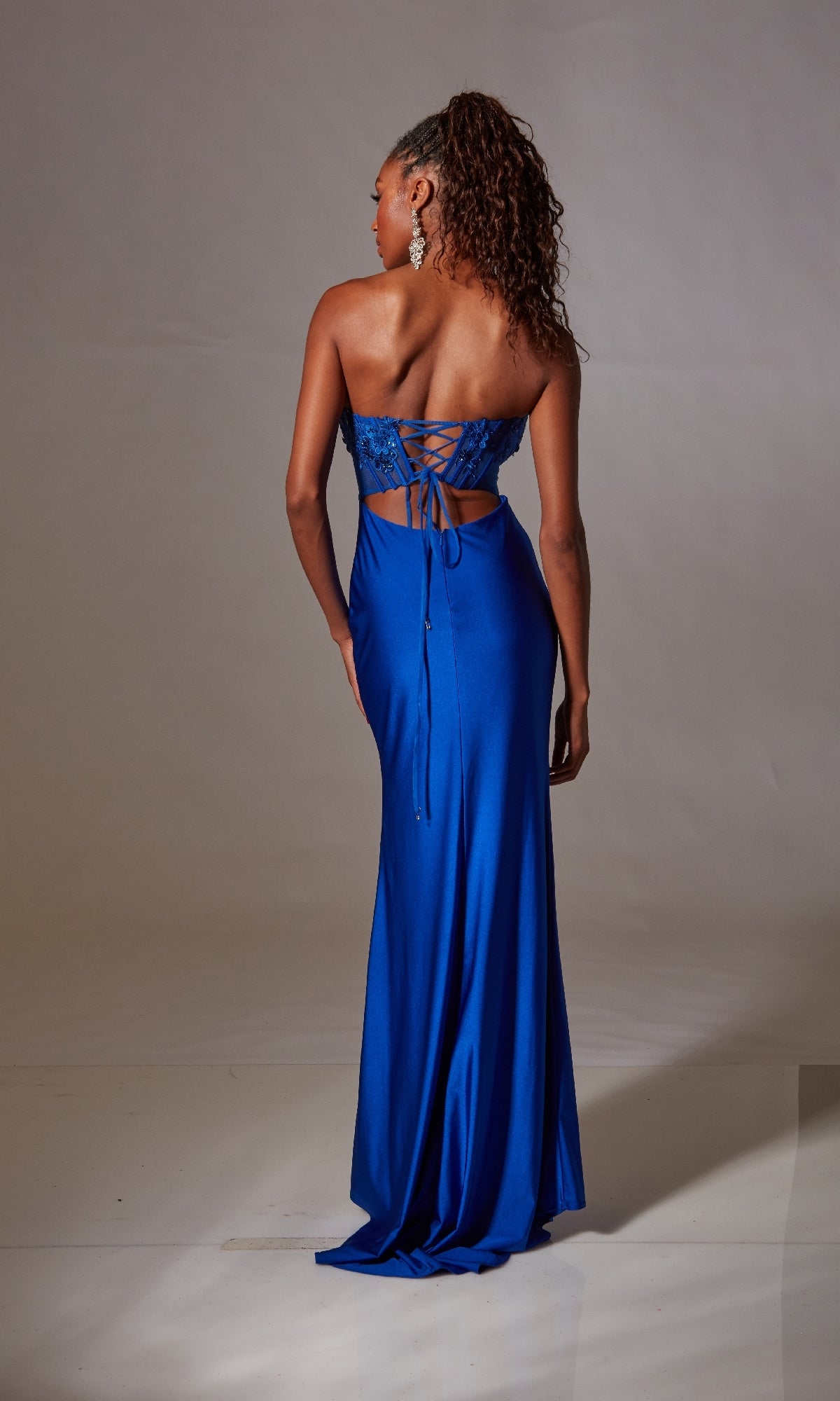 A woman stands facing away, wearing the Blue Strapless Long Prom Dress: Aerie Couture 248, featuring a corset-style bodice, lace-up back detailing, and a small cutout below the ties.
