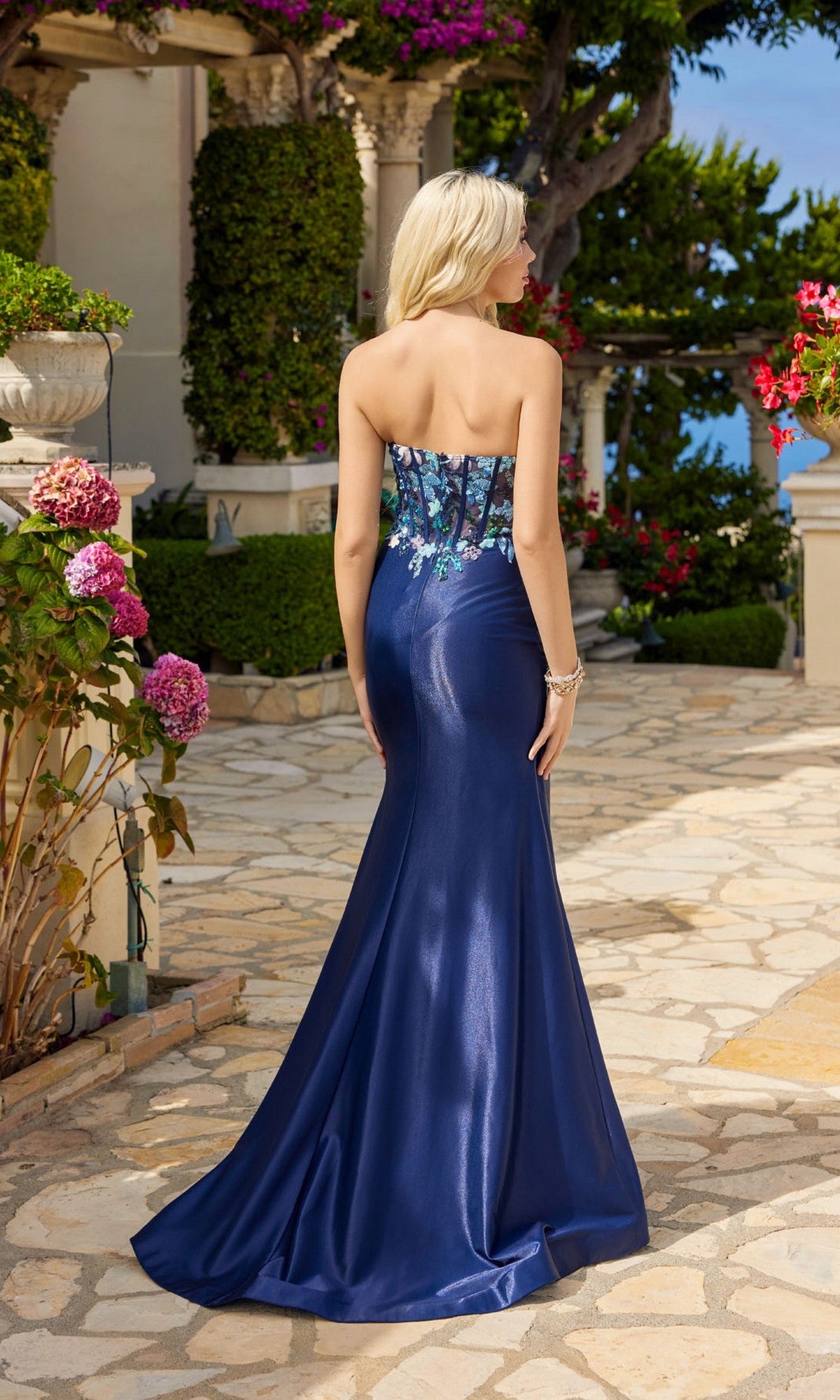 Outdoors in a garden, a woman wears the Strapless Long Blue Prom Dress: Juliet JT2496A, featuring floral embroidery on the bodice for an elegant and enchanting look.