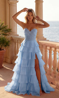 A woman in a light blue Strapless Long Ruffled Prom Gown (PromGirl 250001) with a sheer corset bodice and thigh-high slit stands on a balcony overlooking the ocean.