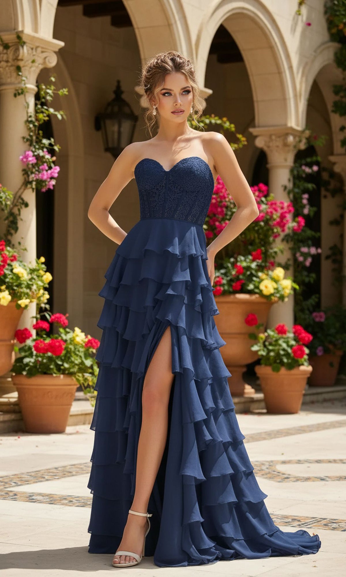 A woman in the PromGirl 250001 Strapless Long Ruffled Prom Gown with a corset-style bodice and thigh-high slit stands outdoors by potted flowers and stone arches.