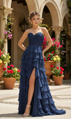 A woman in the PromGirl 250001 Strapless Long Ruffled Prom Gown with a corset-style bodice and thigh-high slit stands outdoors by potted flowers and stone arches.