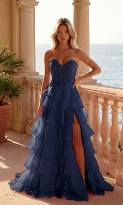 A woman stands on a terrace by the sea wearing the PromGirl 250001 Strapless Long Ruffled Prom Gown in navy, featuring a sheer corset-style bodice and an elegant A-line skirt with a side slit.