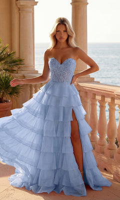 A woman in a periwinkle Strapless Long Ruffled Prom Gown (PromGirl 250001) with a sheer corset-style bodice and thigh-high slit stands on a balcony overlooking the ocean.
