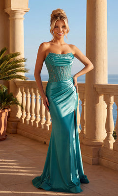 Woman in an emerald Strapless Lace-Bodice Prom Dress (PromGirl Exclusive) with a fitted bodice and draped skirt featuring a high side slit stands on a balcony overlooking the sea, framed by columns and potted plants.