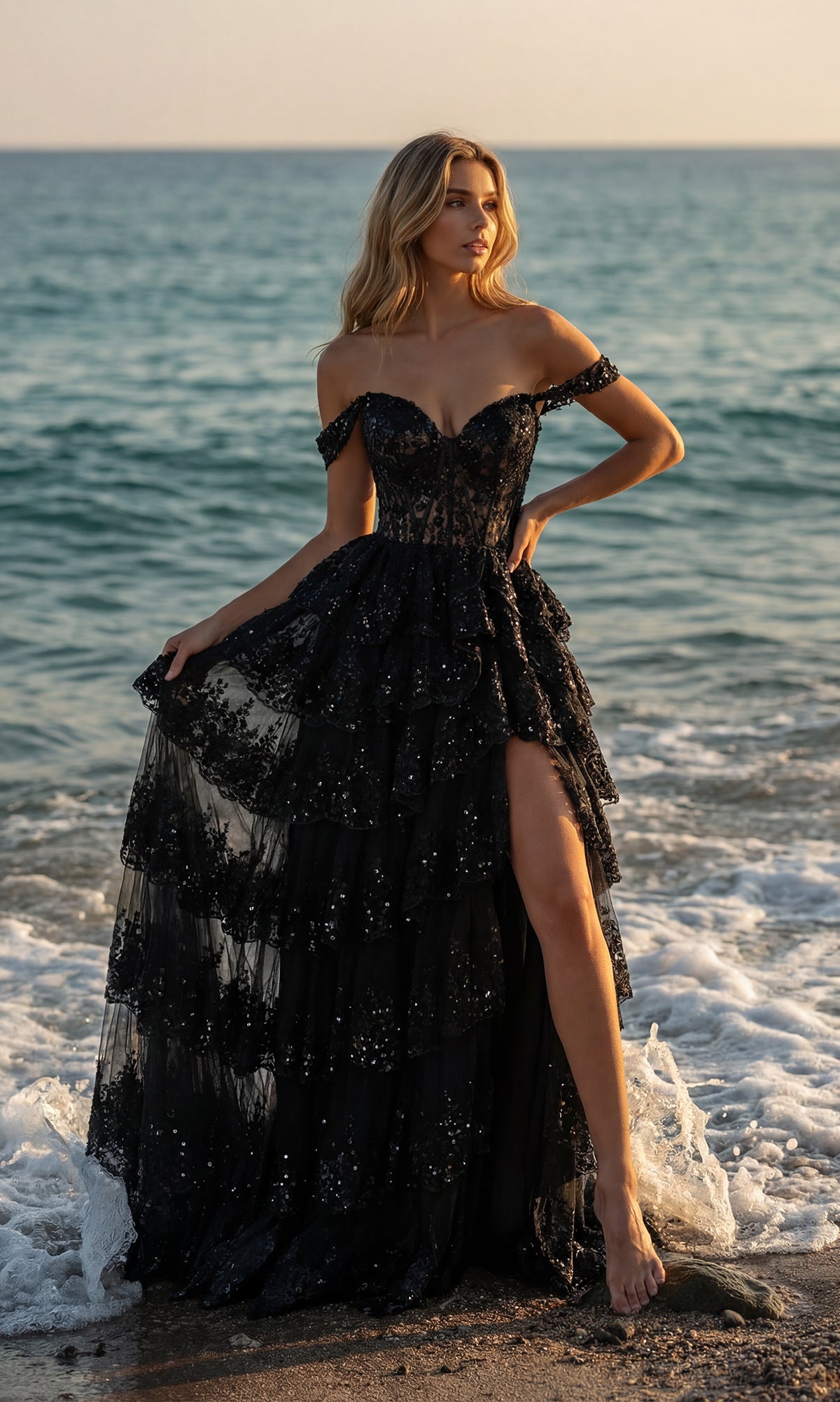 A woman wearing the PromGirl 250005 A-Line Corset Lace Prom Dress in black stands on the shoreline, an off-the-shoulder style with a thigh-high slit and sequins shimmering by the waves.