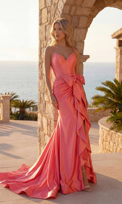 A woman stands outdoors before stone arches, wearing the PromGirl Exclusive 250025 Mermaid Prom Dress in candy pink with bow and ruffle details, as the ocean appears in the background.
