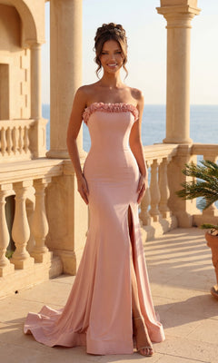 A woman wears the PromGirl Exclusive blush prom dress with mini ruffles, featuring a strapless, floor-length design and front slit, as she stands on a sunlit terrace with stone columns.