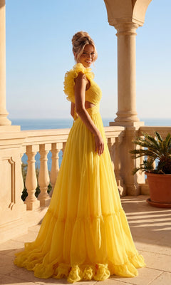 A woman in the PromGirl Exclusive Long Prom Dress with Ruffle Straps stands on a stone balcony overlooking the ocean, near a potted plant beneath a clear sky.