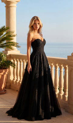A woman in a black Strapless Prom Ball Gown (PromGirl Exclusive 250039) stands on a sunlit terrace with a stone balustrade, overlooking the sea.