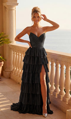 A woman smiles on a balcony overlooking water, wearing the black Strapless Sparkly Prom Dress: PromGirl 250051 with ruffled tiers and a thigh-high slit, one hand on her hip.