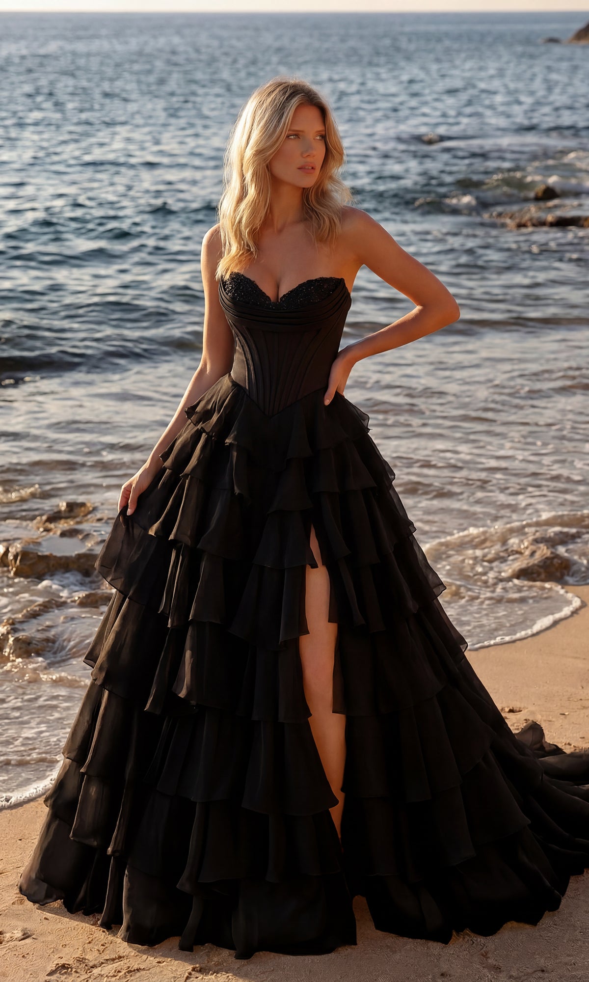 A woman wearing the black Strapless Ruffled Prom Ball Gown by PromGirl 250057 stands on a sandy beach near the shoreline, waves rolling in behind her.