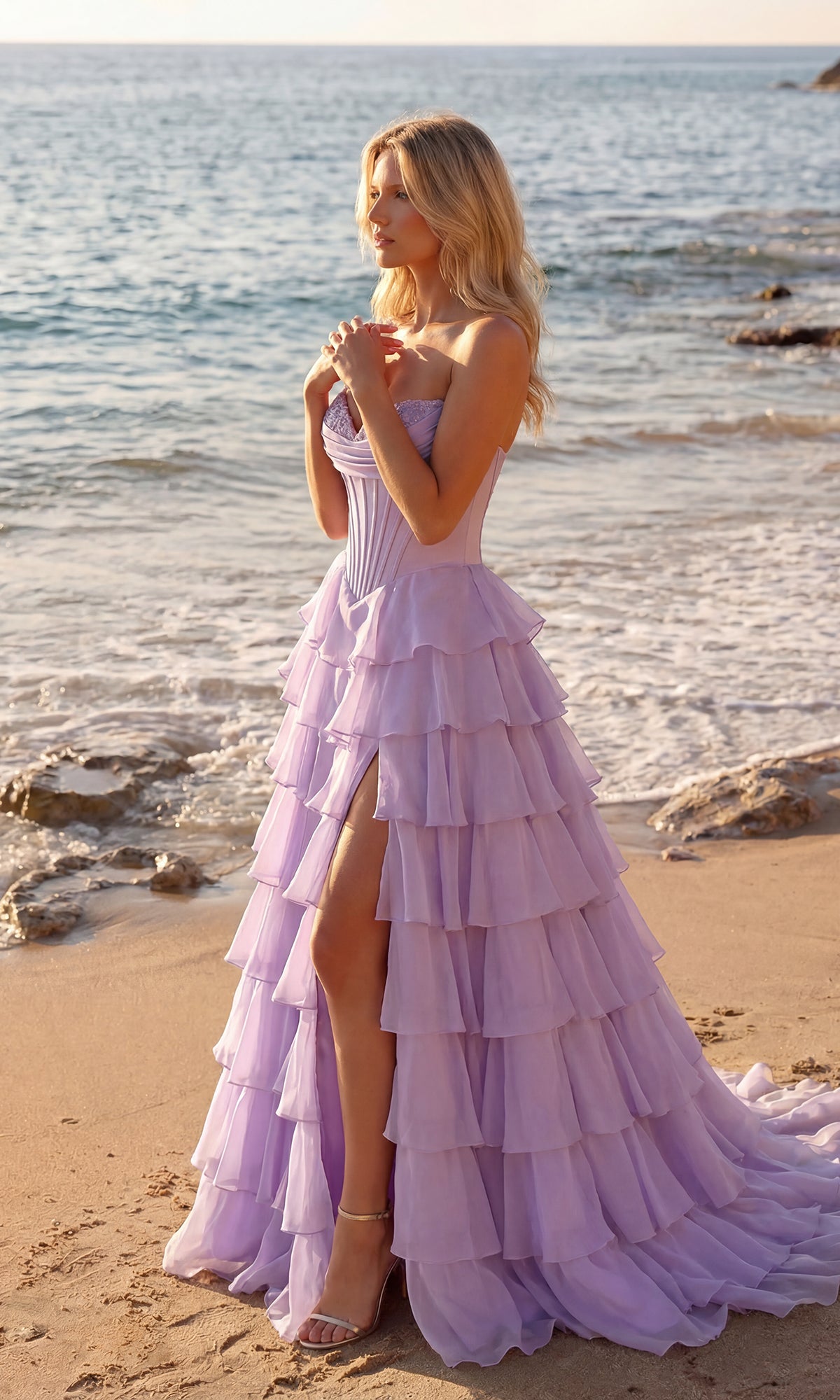 A woman in the PromGirl 250057 Strapless Ruffled Prom Ball Gown stands on a sandy beach near the shoreline, with waves and rocks creating a dreamy backdrop.