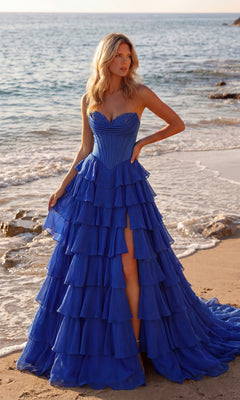 A woman stands on a sandy beach in the PromGirl 250057 Strapless Ruffled Prom Ball Gown, featuring a navy corset bodice, ruffled tiered skirt, and dramatic thigh-high slit.