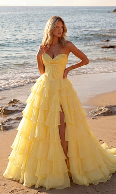 A woman in a light yellow PromGirl 250057 strapless ruffled prom ball gown stands on a sandy beach near the shoreline, with the ocean and rocks behind her.