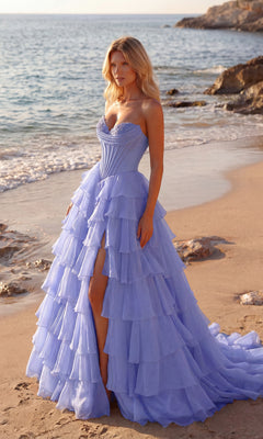 A woman in a periwinkle Strapless Ruffled Prom Ball Gown (PromGirl 250057) stands on a sandy beach near the shoreline, with waves and rocky cliffs in the background.