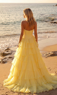 A woman in the PromGirl 250057 Strapless Ruffled Prom Ball Gown with yellow layered tiers stands on a sandy beach facing the ocean, with gentle waves and rocks in the background.
