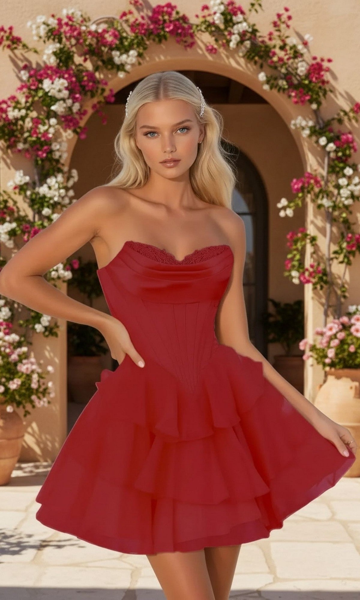 A woman with long blonde hair wears the PromGirl 250092 strapless short homecoming dress with ruffles, posing outdoors by an arched doorway and blooming pink and white flowers.