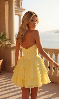 A woman wearing the Strapless Short Homecoming Dress: PromGirl 250092 stands on a balcony overlooking water, sunlight illuminating her layered yellow corset dress.