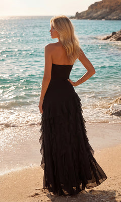 A woman wearing the PromGirl 250104 Ruffled Prom Dress with Slit stands on a sandy beach facing the ocean, with waves and rocky cliffs in the background.