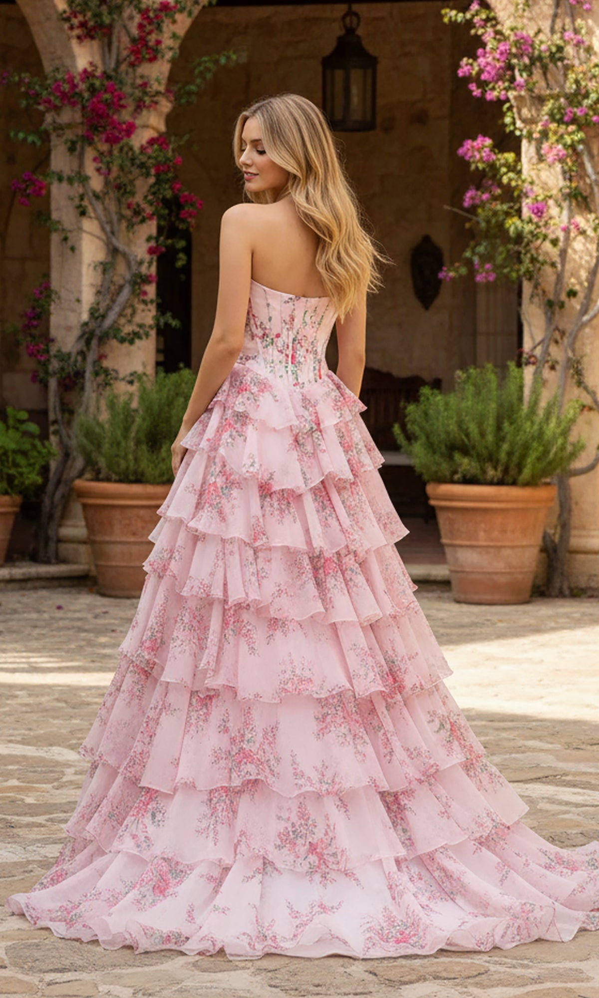A woman with long blonde hair stands outdoors among potted plants and stone arches, wearing the PromGirl Exclusive 250123C long prom dress featuring a strapless floral design and satin corset bodice. She faces away from the camera.