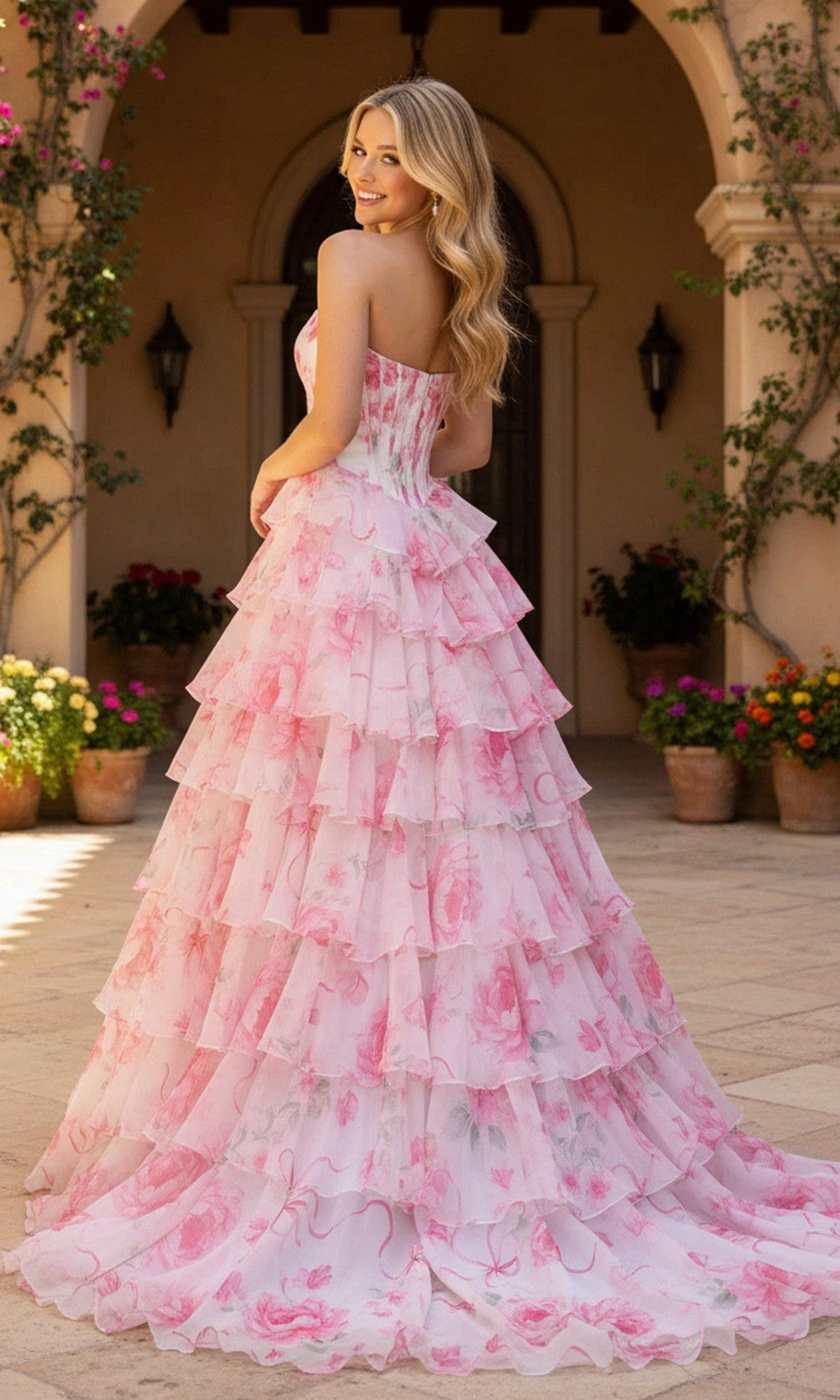 A woman wearing the Long Prom Dress: PromGirl Exclusive 250123D, a strapless floral floor-length pink ruffled gown, smiles over her shoulder on a stone patio with potted plants and an arched doorway in the background.