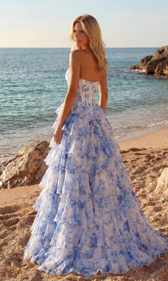 A woman in the Blue Floral Prom Ball Gown: PromGirl Exclusive 250123 stands on a sandy beach by rocks, facing the ocean under a clear sky.