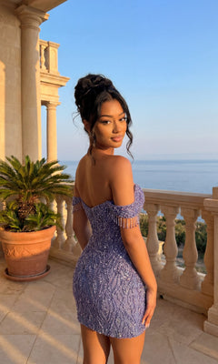 A woman in the Beaded Glitter Hoco Dress: PromGirl Exclusive 250133 with off-the-shoulder sequined sleeves stands on a balcony with columns, overlooking water and greenery under a clear sky.
