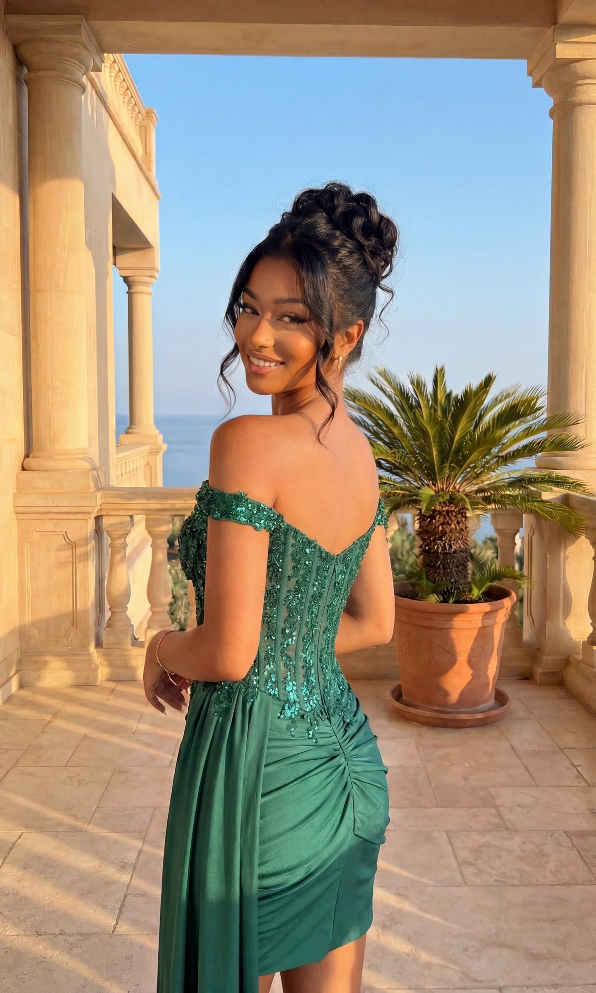 A woman smiles at the camera on a stone balcony with columns, wearing the PromGirl Exclusive 250138 Off-Shoulder Hoco Dress; potted palm and ocean view appear in the background.