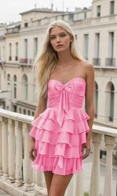 A woman with long blonde hair wears the PromGirl Exclusive 250152 short Hoco dress, a pink strapless homecoming style with ruffles and a front bow, while standing on a balcony overlooking city buildings.