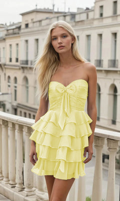 A woman stands on a balcony wearing the PromGirl Exclusive 250152, a strapless, layered yellow a-line short hoco dress with a front bow; buildings appear in the background.