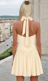 Woman showing the back of a pale yellow short dress with a halter neckline tied in a bow, standing outside.