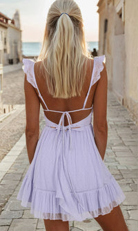 Woman showing the open back of a lilac purple short dress, standing on a cobblestone road